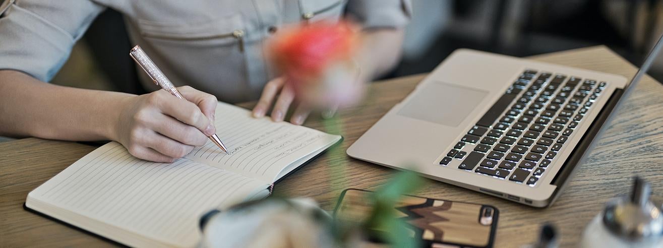 person writing in notebook with laptop